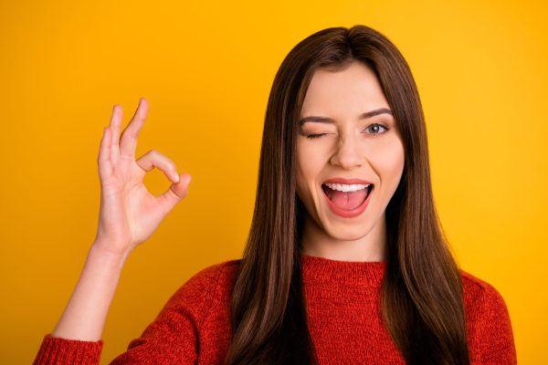 Close up photo of funny funky cheerful girl promoter feel crazy wink blink show, okay sign recommend great ads approve wear red sweater isolated over yellow color background